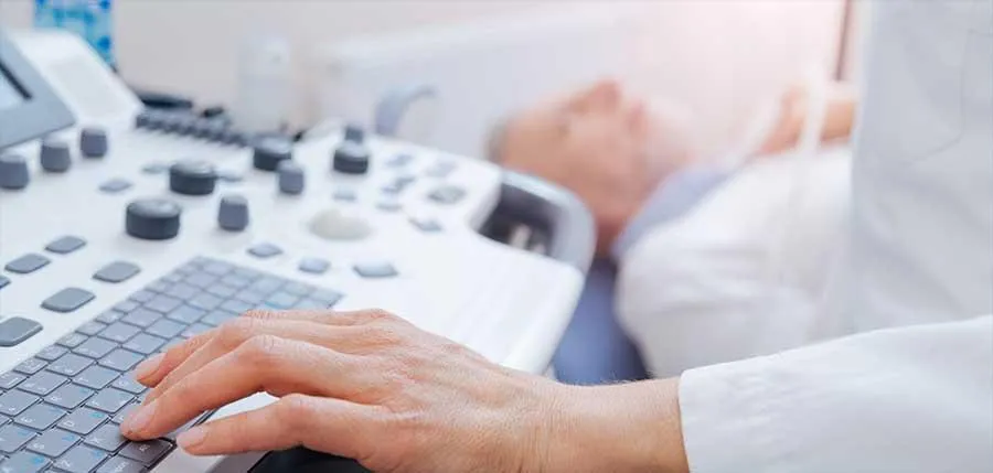 A sonographer is resting his left hand on an ultrasound scanner while performing a thyroid scan on a male patient.